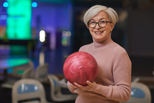 Waist up portrait of smiling senior woman holding bowling ball and looking at camera while enjoying active entertainment at bowling alley, copy space