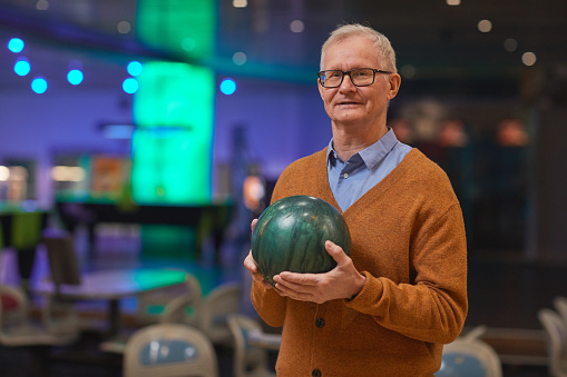 Waist up portrait of smiling senior man holding bowling ball and looking at camera while enjoying active entertainment at bowling alley, copy space