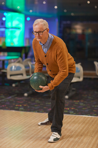 Vertical full length portrait of active senior man playing bowling, standing by lane ready to throw while enjoying entertainment