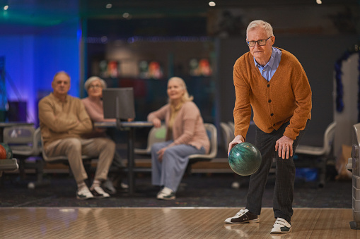 Full length portrait of active senior man playing bowling with group of friends in background, copy space