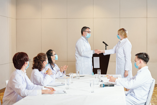 Experienced researchers in medical masks shaking hands after having successful medical conference dedicated to coronavirus