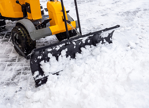 A large orange tractor removes snow from the road and clears the sidewalk. Cleaning and clearing roads in the city from snow in winter. Snow removal after snowfalls and blizzards.