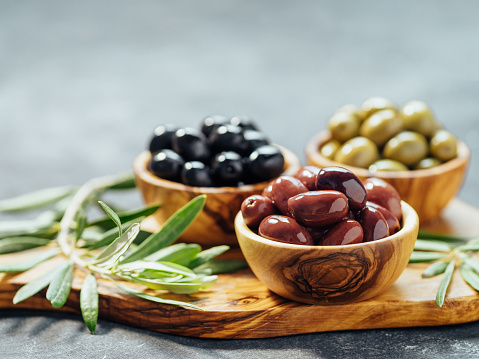 Set of green, red and black olives on gray background. Different types of olives in olive wooden bowls and olive oil over wooden cutting board and fresh olive leaves. Copy space.