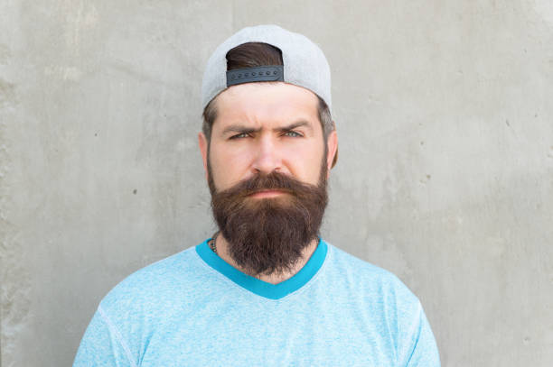 Bearded and moustached. Bearded man on grey wall. Serious bearded hipster in casual summer style. Caucasian guy wearing long mustache and beard hair on bearded face.