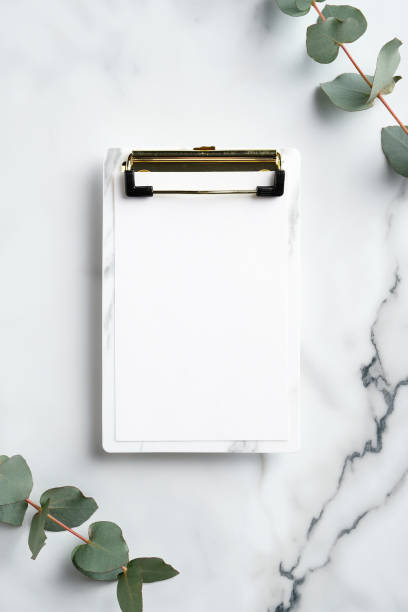 Clipboard with blank paper and eucalyptus branches on marble desk. Flat lay, top view. Wedding checklist concept.