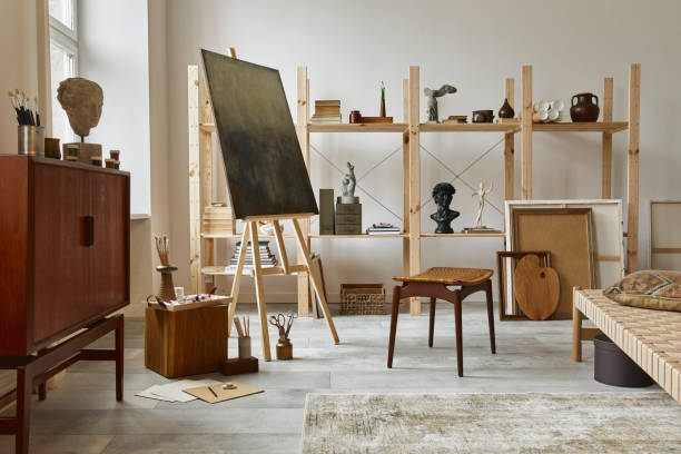 Unique artist workspace interior with stylish teak commode, wooden easel, bookcase, artworks, painting accessories, decoration and elegant personal stuff. Modern work room for artist. Template.