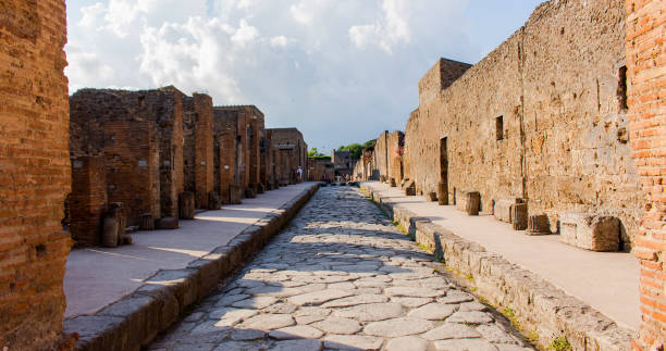 Ancient paved street in the ruins of Pompeii, Italy Ancient paved street in the ruins of Pompeii, Italy. Roman town destroyed and buried by ash during the eruption of Vesuvius volcano. A cloudy summer day with tourists wandering in the back. – Image pompeii ruins stock pictures, royalty-free photos & images