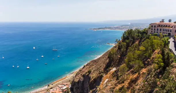 Panoramic landscape of Taormina a sunny summer day in Sicily (Italy). Sicilian seascape, beach and blue sky, with luxury hotel and green vegetation in the foreground. – Image