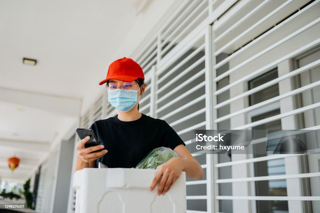 An Asian Chinese woman using smartphone and delivering fresh vegetables to customer Image of an Asian female delivery person with protective face mask sending fresh vegetables to a customer. She is using smartphone to contact the customer or check orders. 30-34 Years Stock Photo