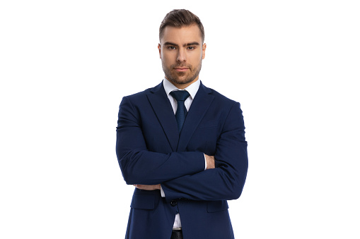 serious young man in navy blue suit folding arms and posing on white background in studio, portrait