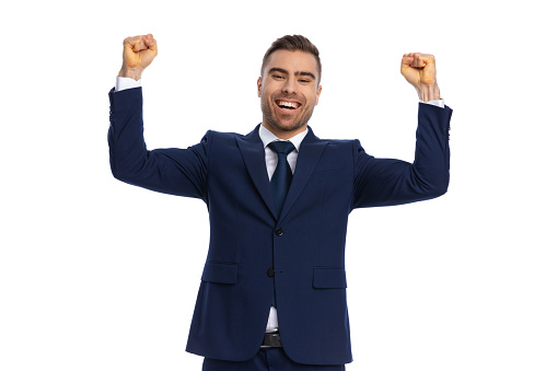excited young businessman in navy blue suit holding fists in the air, laughing and celebrating victory, standing and posing isolated on white background in studio, portrait