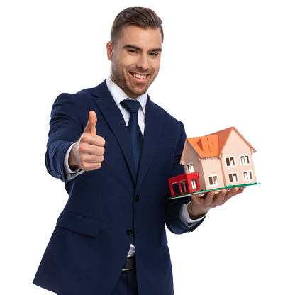 confident real estate agent in navy blue suit posing isolated on white background, making thumbs up gesture and smiling, showing and presenting building model in studio