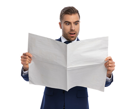 astounded young guy in elegant navy blue suit reading newspaper in a shocked manner, standing isolated on white background in studio