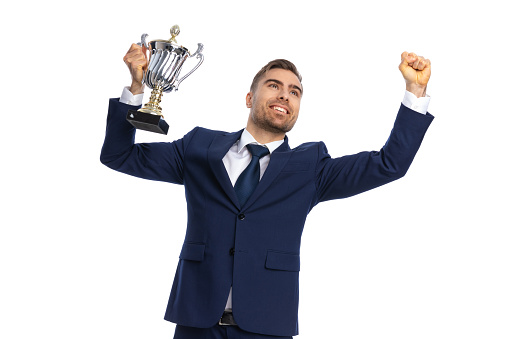 excited young guy in navy blue suit looking up, holding fists and trophy in the air and celebrating victory on white background in studio