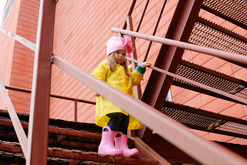 A blonde girl in a yellow raincoat and pink rubber boots comes down the stairs