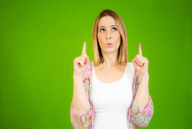 istock Young woman pointing up with both index fingers while looking up, standing over green background. 1298312298