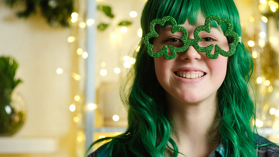 Joyful teen girl with green hair smiling wearing funny glasses in shape of clover celebrating Saint Patrick's day.