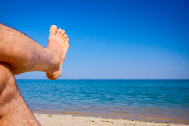 Man's crossed legs are sunbathing by lying carefree on sand next to the coastline, on public beach.