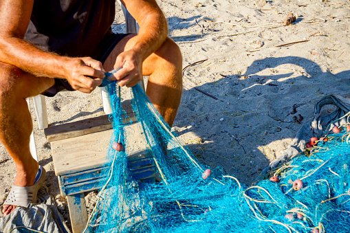 A fisherman sits on a chair at the beach, piles up fishing net and cleans it from shells.