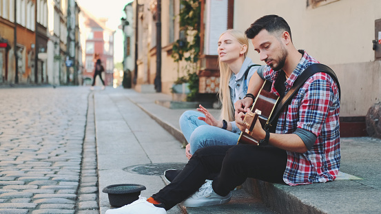 Young couple of street singers sitting on sidewalk, playing guitar and singing. Lifestyle concept.