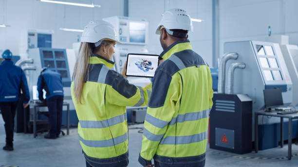 istock Industry 4.0 Factory: Chief Engineer and Project Supervisor in Safety Vests and Hard Hats, Talk, Use Digital Tablet Computer, Screen Shows 3D Concept of New Engine. Industrial Workshop with Machinery 1298326692