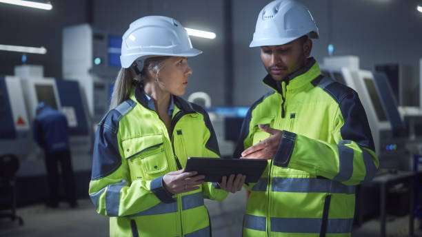 istock Chief Engineer and Project Manager Wearing Safety Vests and Hard Hats, Use Digital Tablet Computer in Modern Factory, Talking, Optimizing Production Line. Industrial Facility with CNC Machinery 1298326696