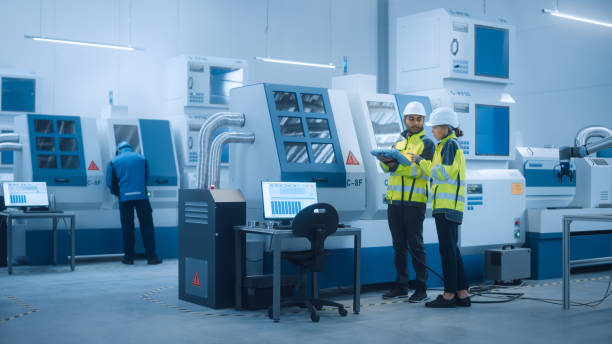 istock Chief Engineer and Project Manager Wearing Safety Vests and Hard Hats, Use Digital Tablet Controller in Factory, Optimizing CNC Machinery Programming Machine for Increasing Production Line Efficiency 1298326799