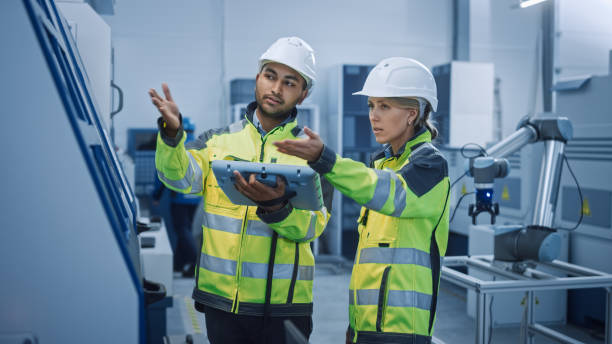 istock Chief Engineer and Project Manager Wearing Safety Vests and Hard Hats, Use Digital Tablet Controller in Modern Factory, Talking, Optimizing CNC Machinery, Programming Machine for Increasing Efficiency 1298327330
