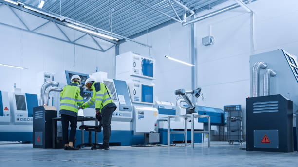 istock Chief Engineer and Project Manager Wearing Safety Vests and Hard Hats, Use Digital Tablet Controller in Factory, Optimizing CNC Machinery,Programming Machine for Increasing Production Line Efficiency 1298327348