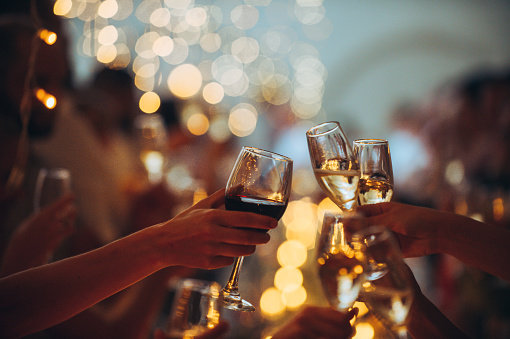 Numerous hands holding champagne flutes and wine glass with red wine with celebratory toast selective focus against string light background