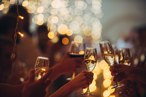 Numerous hands holding champagne flutes and wine glass with red wine with celebratory toast selective focus against string light background