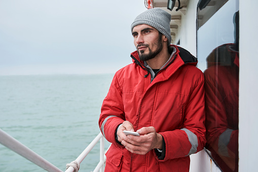 Portrait of a pondering unshaven man thinking about something while holding his smartphone. Man standing at the deck of the boat and waiting for the fish catch. Occupation and technology concept