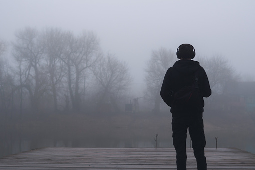 A young man stands alone on a wooden bridge by the lake. Early cloudy foggy morning.