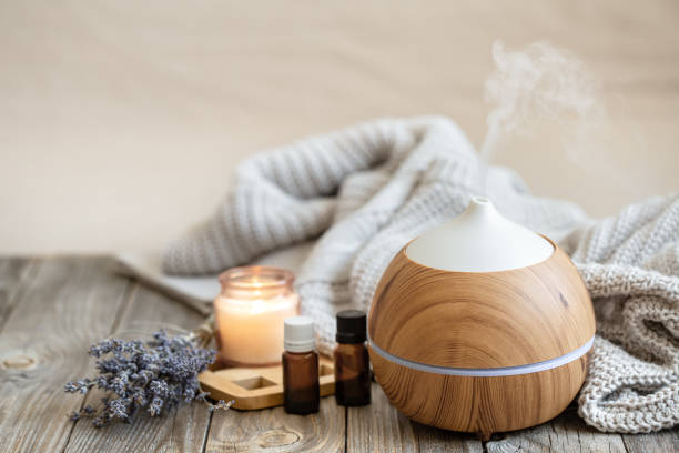 Cozy composition with an air humidifier, a set of aromatic oils and a candle. Modern aroma oil diffuser on wood surface with knitted element, candle and lavender oil on a blurred background. home diffuser stock pictures, royalty-free photos & images