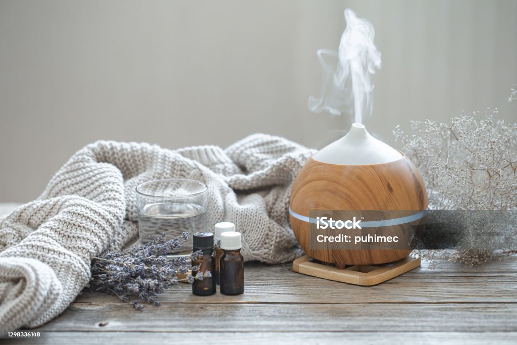 Composition with an air humidifier and a set of aromatic oils. Modern aroma oil diffuser on wood surface with knitted element, water and oils in jars. Aromatherapy Diffuser Stock Photo