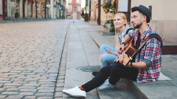 istock Mman and woman sitting on sidewalk, playing guitar and having rest 1298338415
