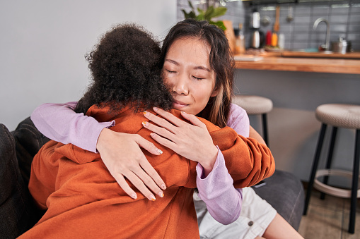 Two sad diverse women talking at home. Female friends supporting each other. Portrait view of the woman embracing with tenderness her friend. Problems, friendship and care concept