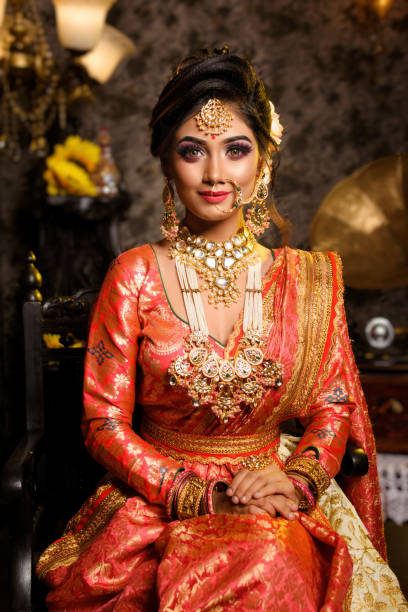 Magnificent young Indian bride in luxurious bridal costume with makeup and heavy jewellery is sitting in a chair in with classic vintage interior in studio lighting. Wedding fashion. stock photo