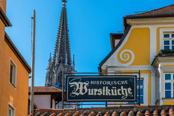 Regensburg, Germany - August 19, 2020: Historic Wurstkuchl in Regensburg / Germany. You get here the traditionel Regensburger food - grilled sausages on sauerkraut with a bun. In background the cathedral of Regensburg.