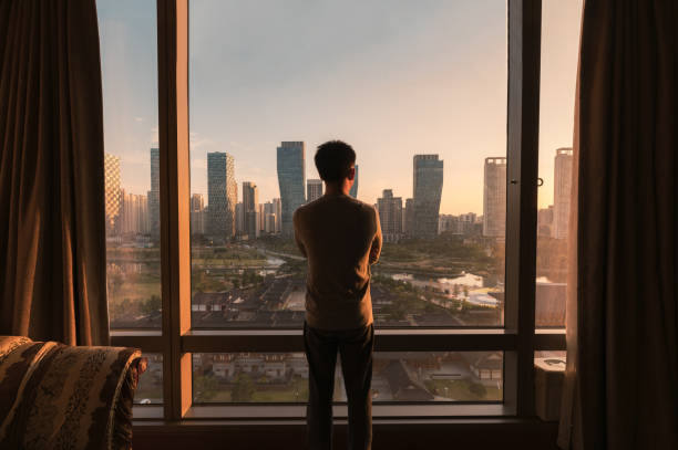 Young asian man standing and looking through window in a hotel. Sunrise over modern buildings in Songdo central park Young asian man standing and looking through window in a hotel. Sunrise over modern buildings in Songdo central park at Incheon, South Korea vista posterior stock pictures, royalty-free photos & images