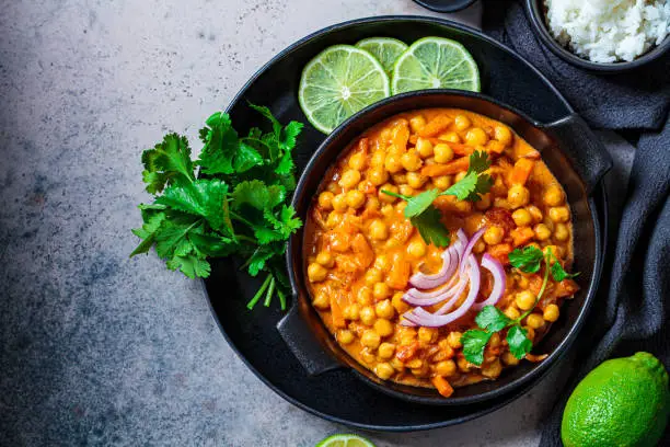 Photo of Vegan chickpea curry with rice and cilantro in black plate, dark background. Indian cuisine concept.