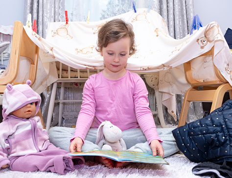 A preschooler child, a girl of 5 years old, built a tent from chairs and sheets. The girl is reading a book to her toys. Children's imagination and creativity at home.