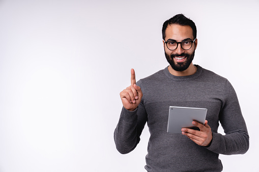 Attractive young Middle Eastern man pointing at copy space with tablet in casual attire isolated over white background
