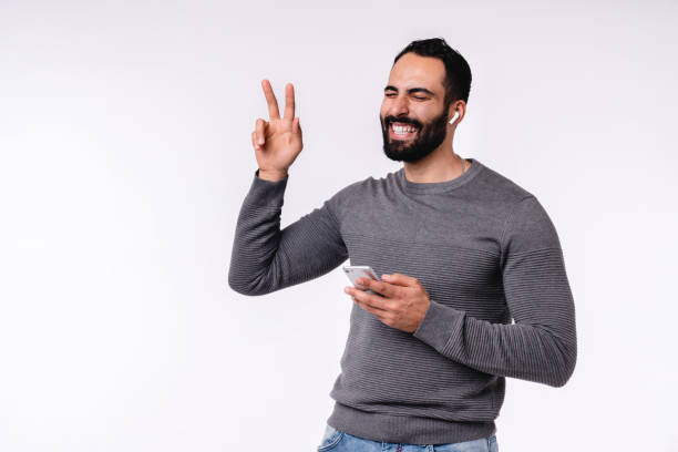 istock Happy cheerful young Arab man showing victory gesture while using smart phone isolated over grey background 1298353692