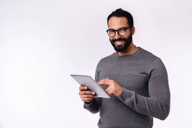 foto de vista lateral de jovem homem árabe bonito usando tablet em roupa casual isolado sobre fundo branco - ipad people holding young adult - fotografias e filmes do acervo