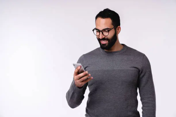 Photo of Nice-looking young Arabian man in casual clothes using mobile phone isolated over white background