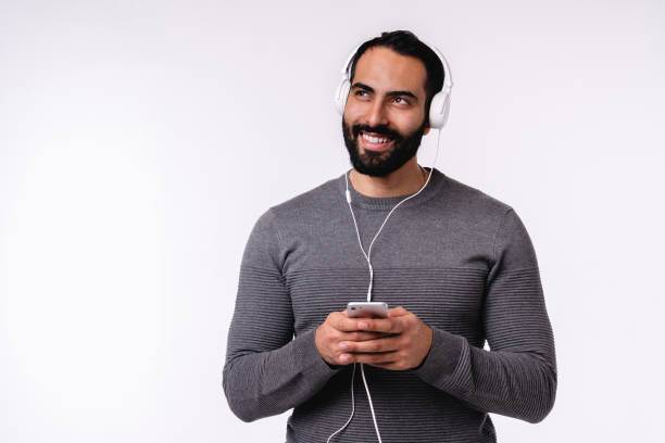 白い背景の上に隔離された彼のスマートフォンとイヤホンで音楽を聴く夢のような若い格好良いアラブ人男性 - young adult technology beautiful singing ストックフォトと画像