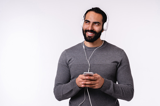 Dreamy young good-looking Arab man listening to the music on his smart phone and earphones isolated over white background