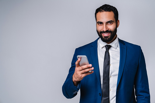 Portrait of a young smiling middle eastern man in business suit using mobile phone isolated over grey background
