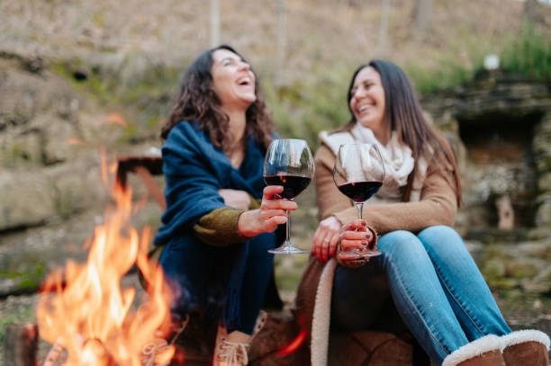 mujeres riendo, sosteniendo copa de vino tinto. las hembras calentando junto al fuego. - outdoors drinking women friendship fotografías e imágenes de stock